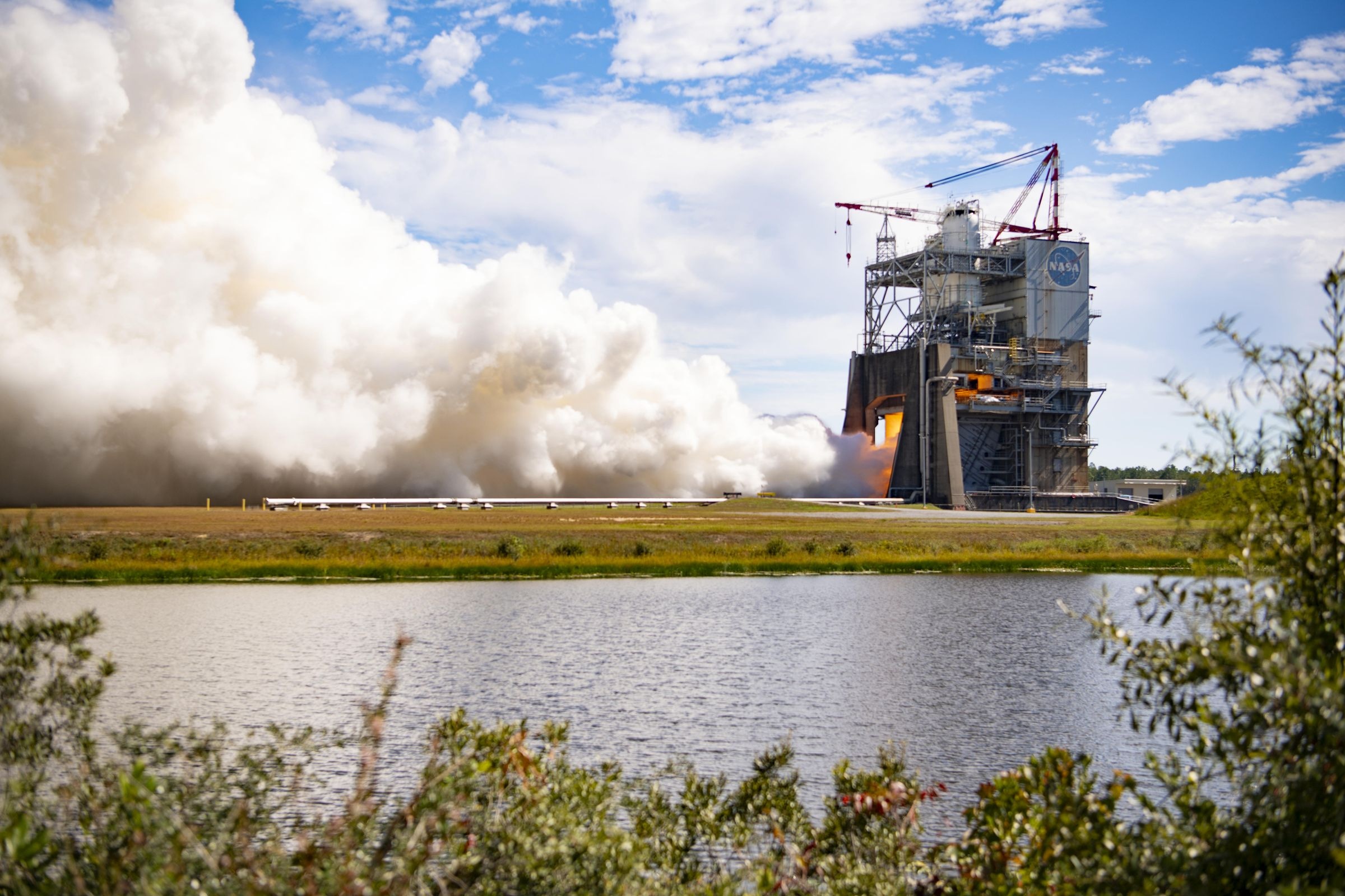 Ракетный двигатель RS-25, закреплённый на испытательном стенде, во время огневого прогона: из сопла бьёт яркая струя пламени.