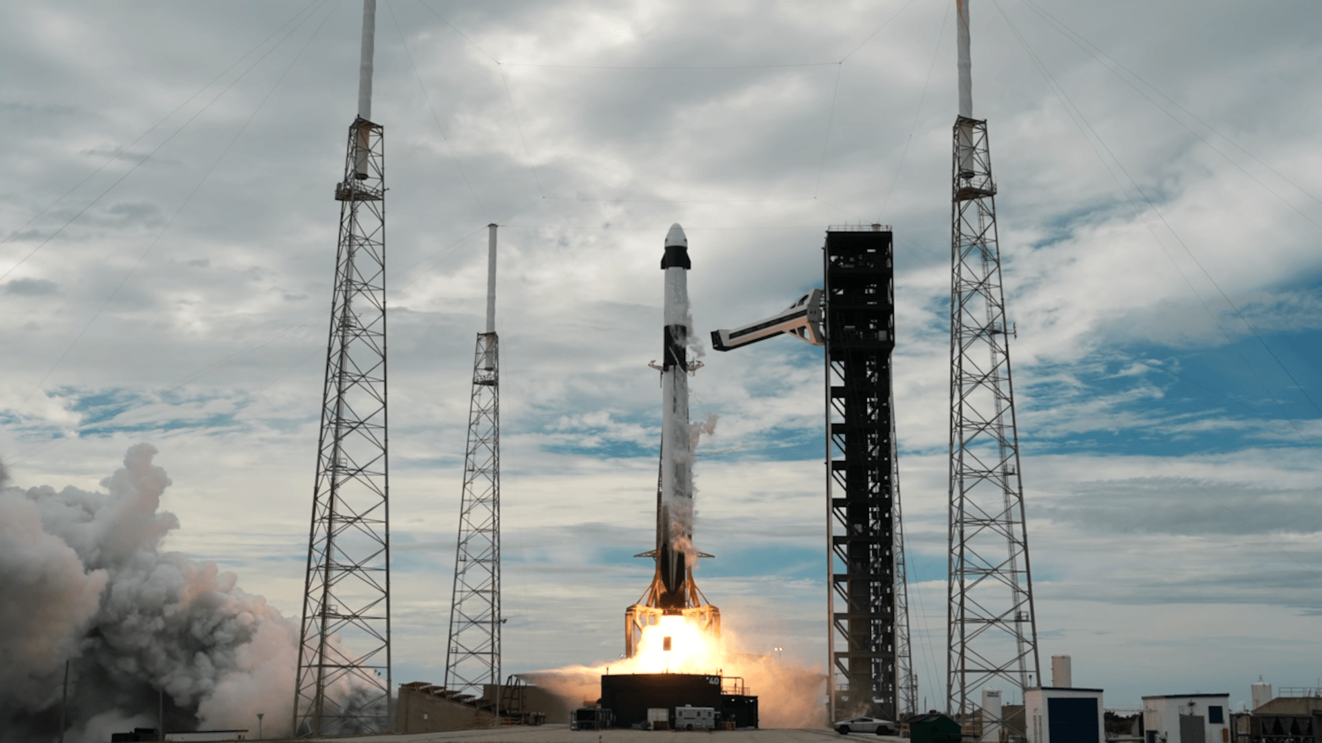 Ракета Falcon 9 с кораблём Dragon на вершине поднимается со стартового комплекса во Флориде. Под ракетой столб пламени и клубы дыма, над ней голубое небо.