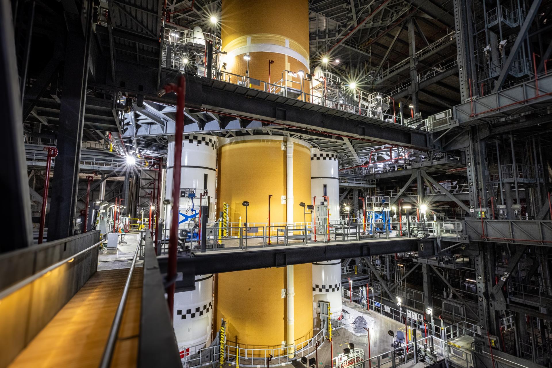 Огромную оранжевую центральную ступень ракеты SLS поднимают внутри высокого сборочного здания, вокруг видны строительные площадки и подъёмное оборудование.